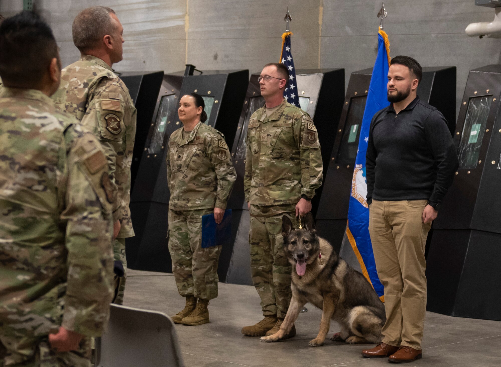 Military working dog, Morgen sits while two uniformed Air Force personnel and one civilian personnel stands at attention.