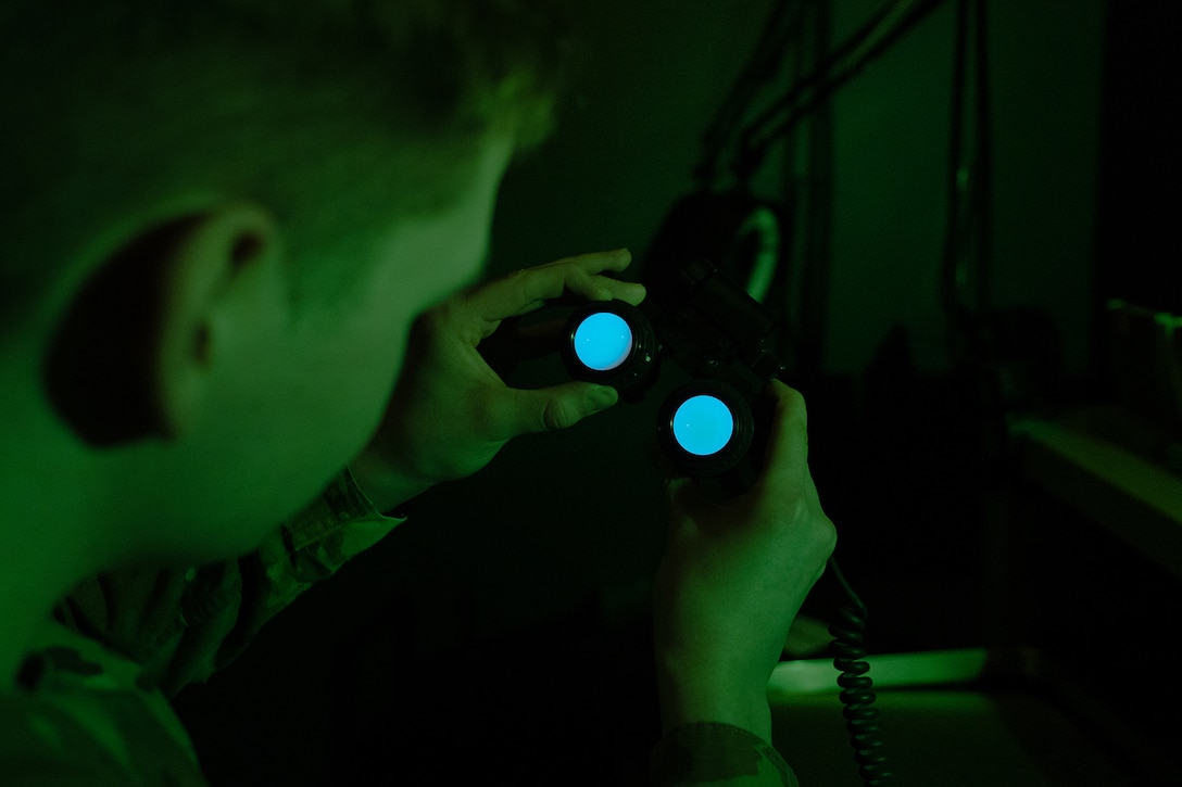 An airman is photographed from the side, at night, inspecting night vision goggles.