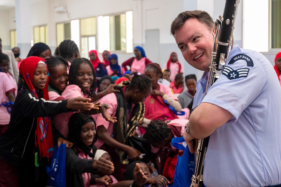 An airman holding a clarinet reaches into a bag in front of a group of children.