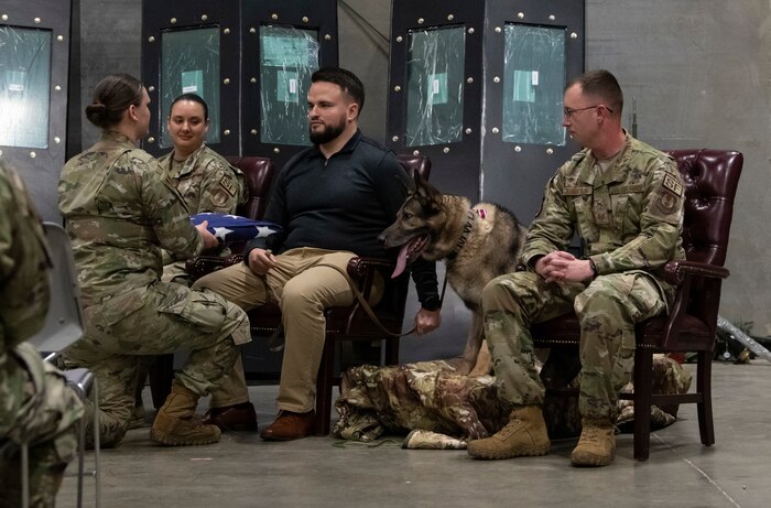 Military working dog, Morgen sits while two uniformed Air Force personnel and one civilian personnel stands at attention.