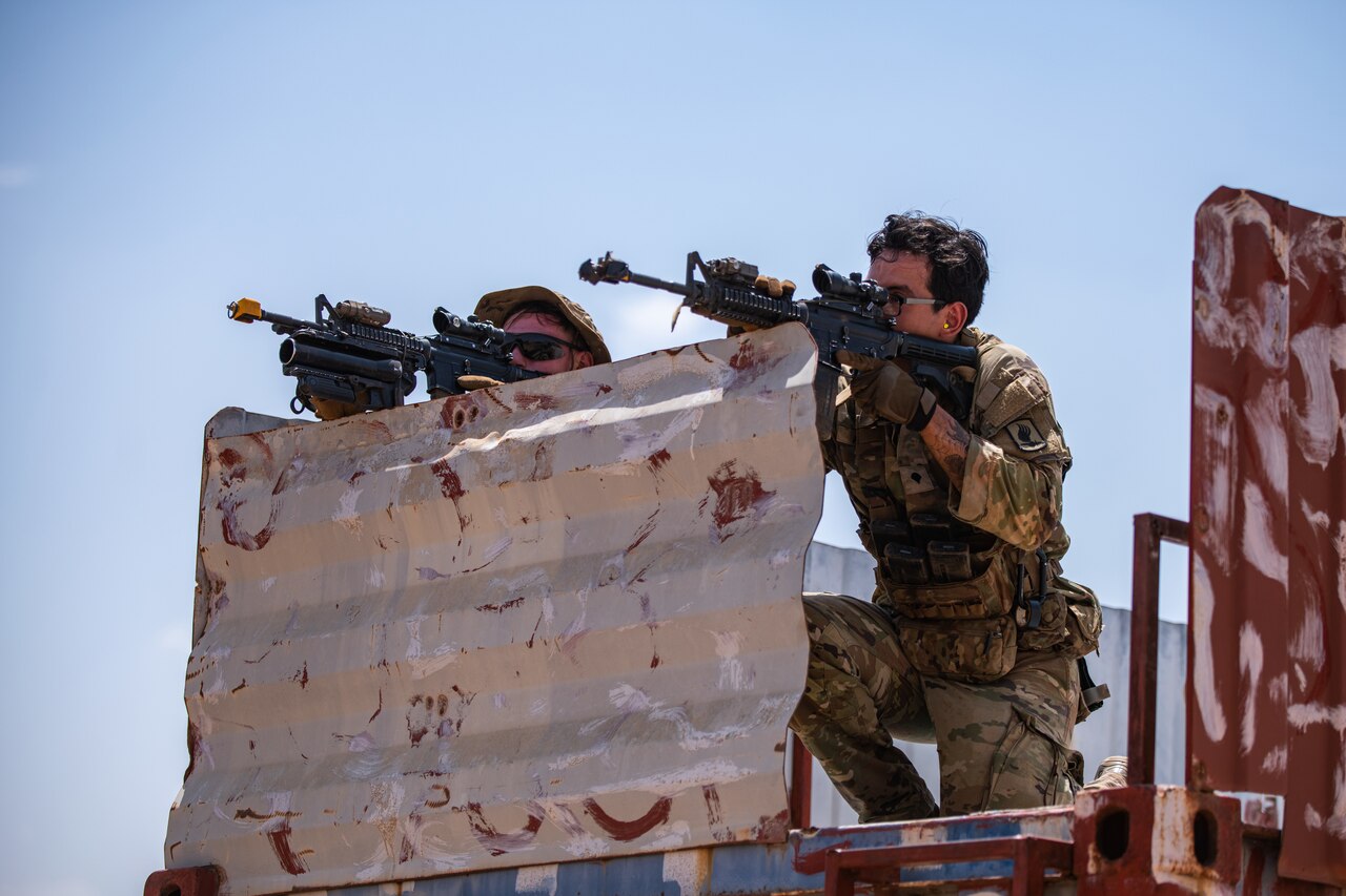 Two soldiers knell behind a barrier and fire their weapons.