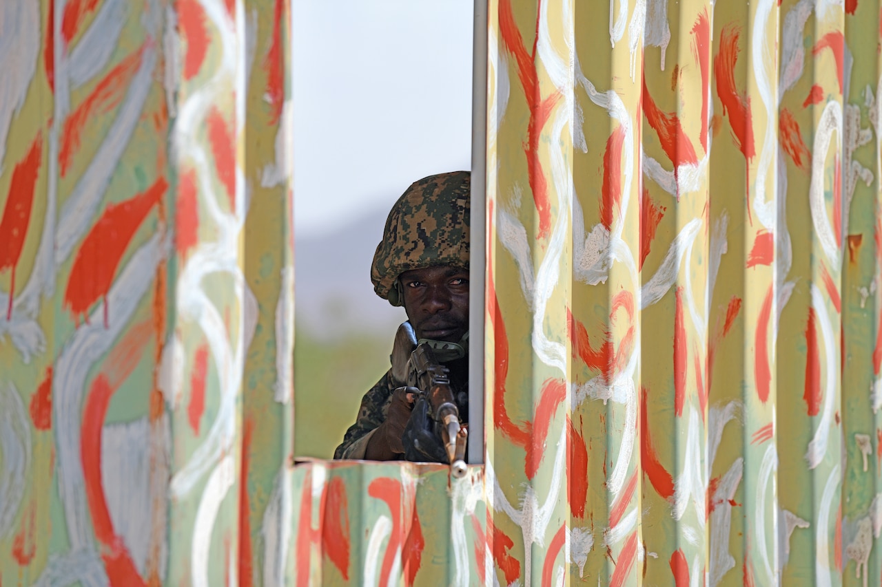 A soldier peers from behind a barrier holding a gun.