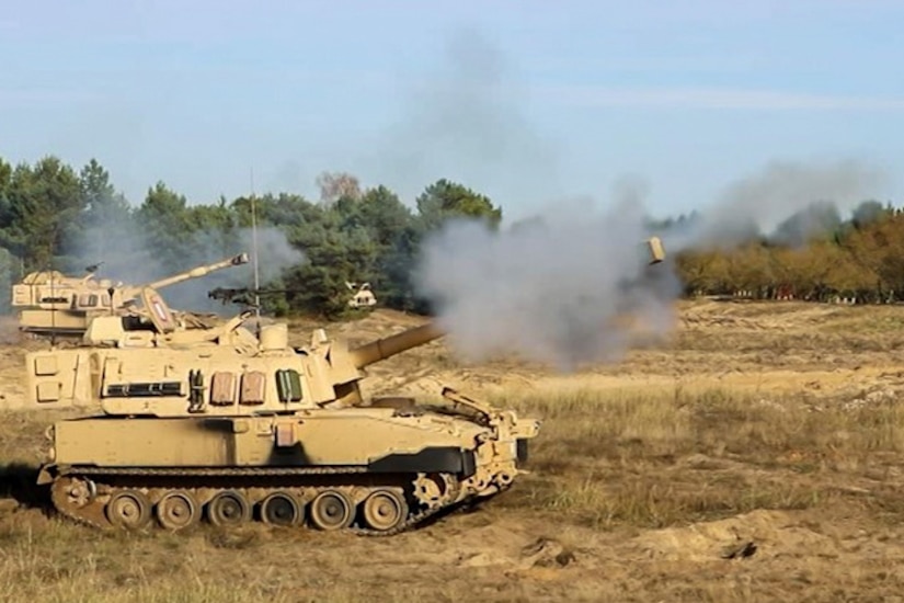 Weapons mounted on a pair of military vehicles fire rounds of ammunition in an open field.