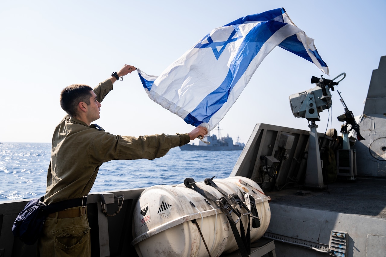 A uniformed service member handles an Israeli flag while standing on the deck of a naval vessel. The flag waves in the wind.