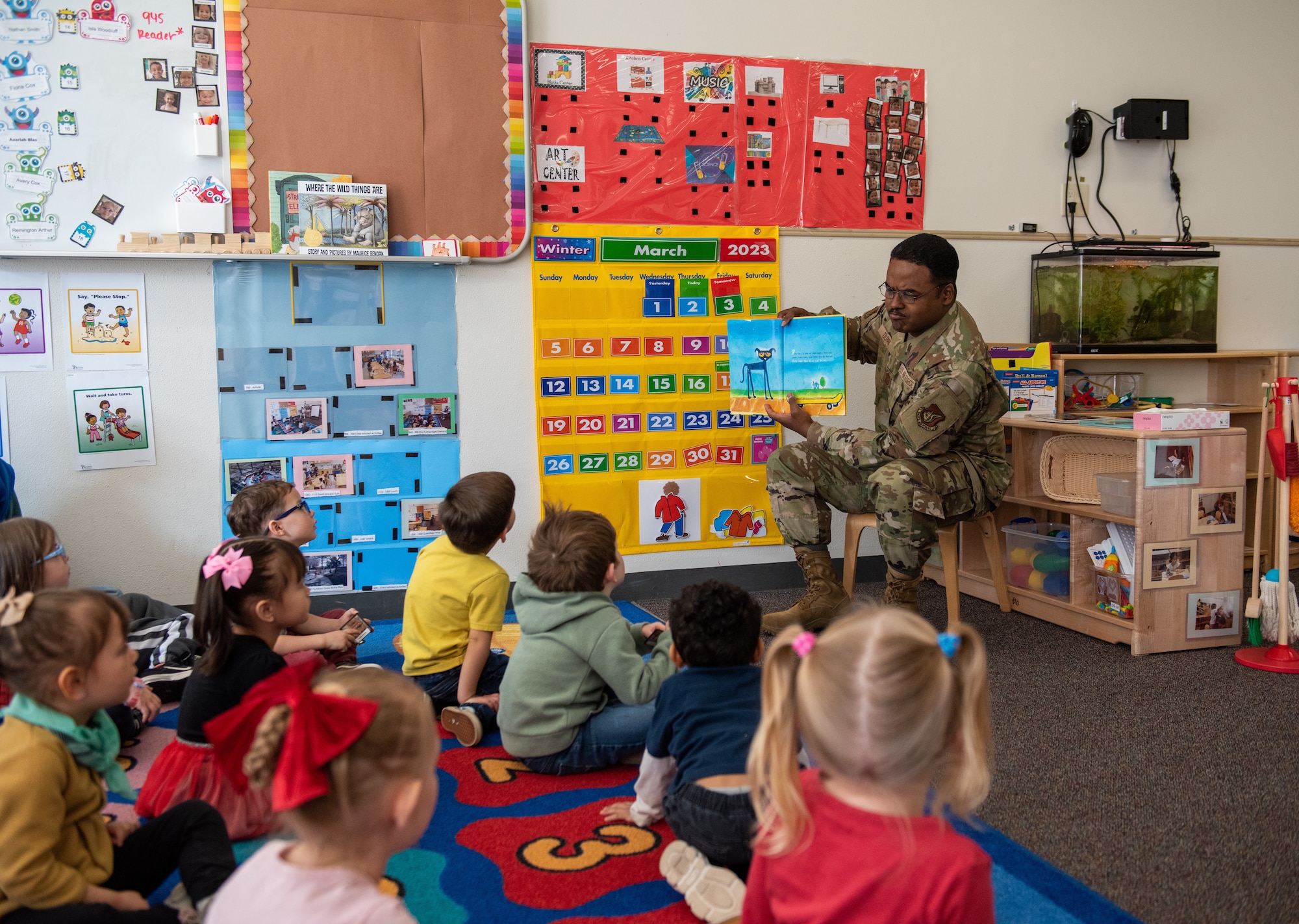 A military member sits in a chair reading to a group of children sitting on the floor.