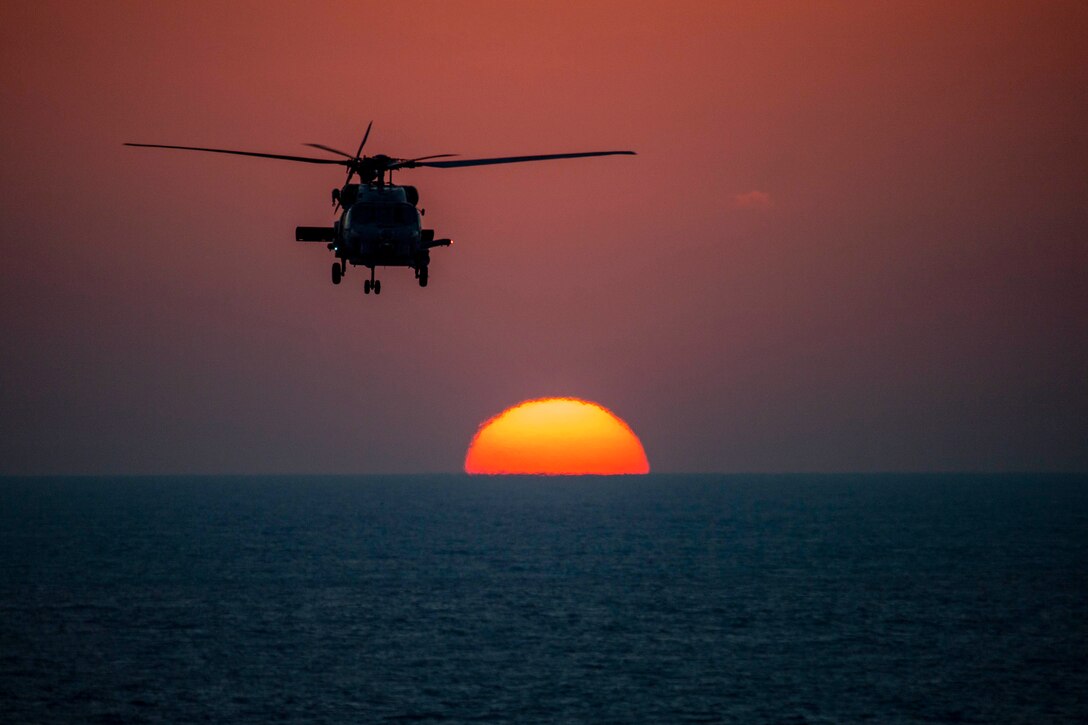 A helicopter prepares to land on an aircraft carrier with the sun in the background.