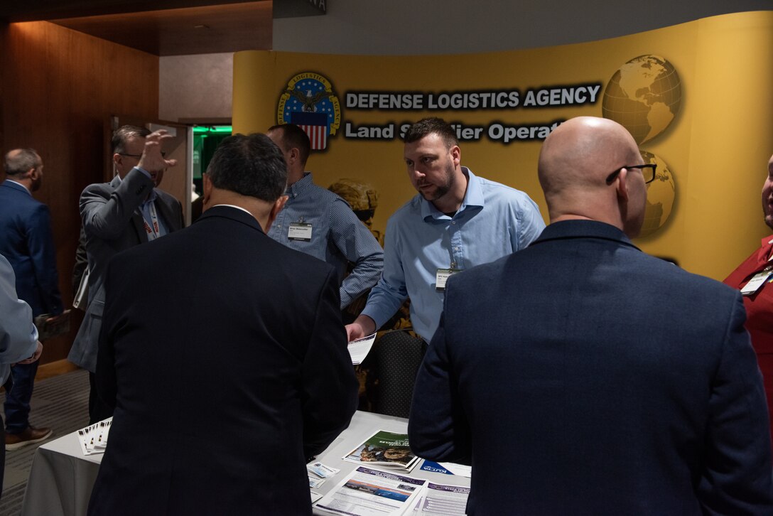 Men in suits visit a DLA Land and Maritime booth with a yellow background in the hallway outside a ballroom.