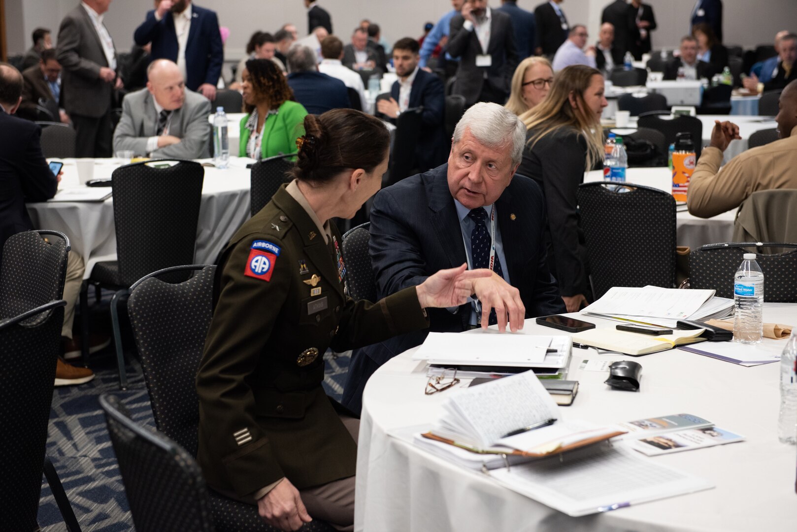A white haired man in a business suit chats with a dark haired woman in an Army dress uniform (olive green coat and tan pants) at a table with a white tablecloth in a ballroom.