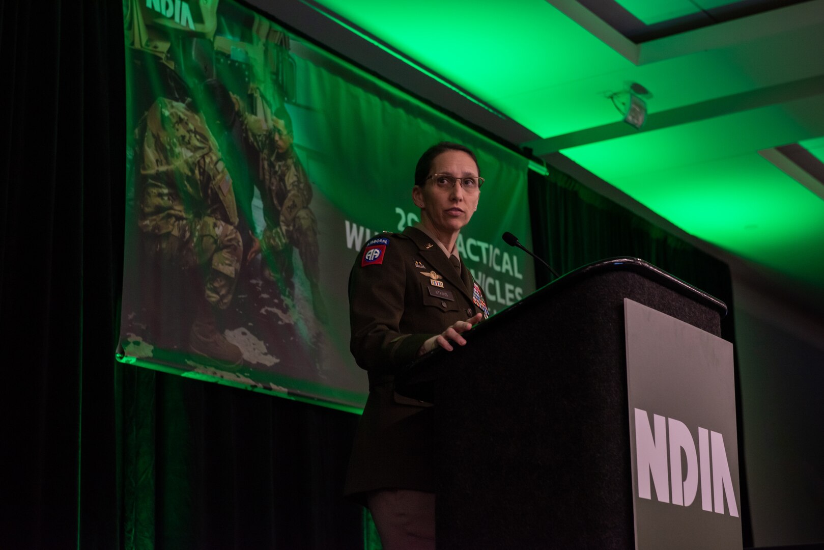 A dark haired woman in an Army dress uniform (green jacket and tan pants) speaks at a podium under green lighting on a stage in a ballroom.