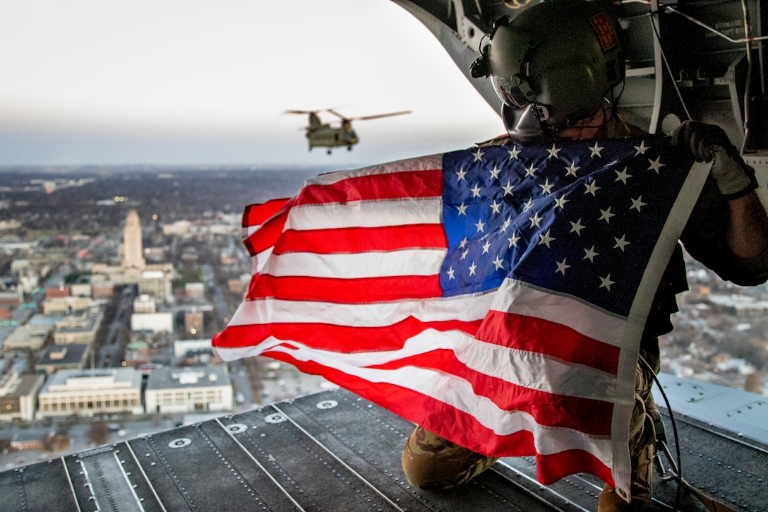 A Nebraska Army National Guard Soldier with Company B, 2-135th General Support Aviation Battalion, holds a U.S. flag on the back of CH-47 Chinook helicopter while flying over Lincoln, Nebraska, where the Capitol Building can be viewed to the left.