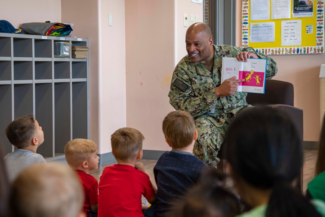 A sailor sits in front of a group of children while reading a book.