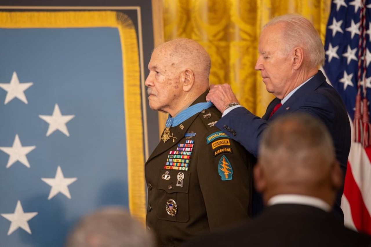 A man places a blue military ribbon and medal around another man’s neck.