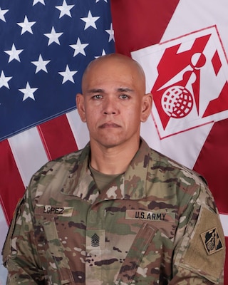 CSM Jaime Lopez, who is wearing the Army OCP uniform. Behind him are the U.S. National Colors and POD Colors.