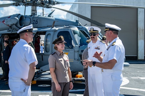 A woman in military uniform speaks to three men in military uniform. A helicopter is in the background.