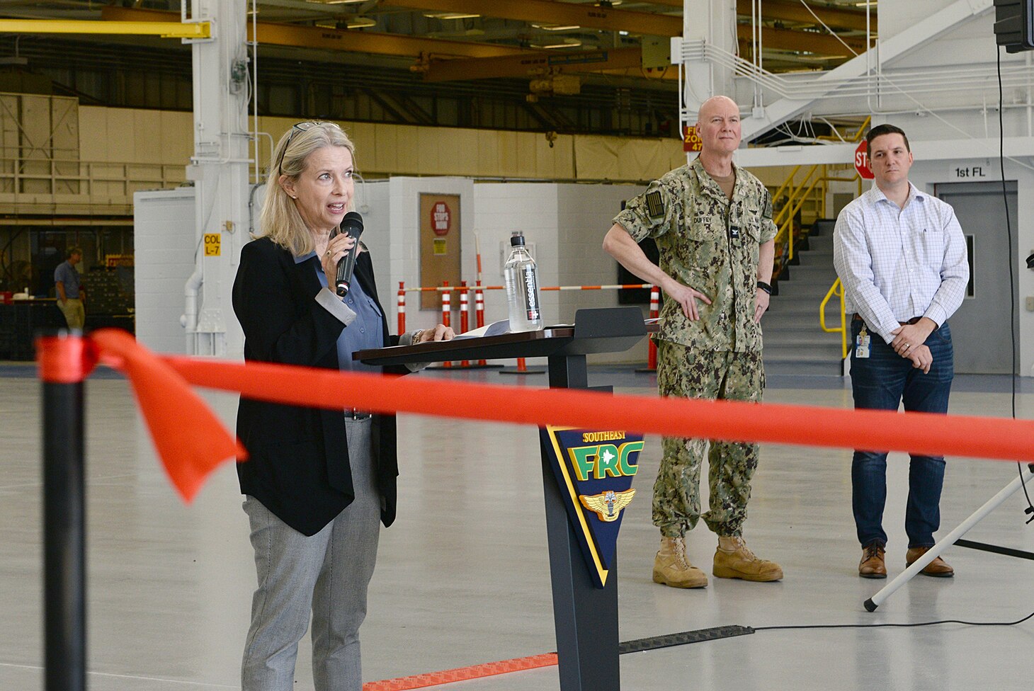 Stacey Barton, Vice President and Division Leader of Haskell Company, Fleet Readiness Center Southeast's (FRCSE) partner in its recent Hangar 101 renovation project, speaks to an audience of spectators at a ribbon-cutting ceremony as FRCSE's Commanding Officer, Capt. Grady Duffey and FRCSE's Facility and Infrastructure Management Department Director, Daniel Simon, look on. The renovation upgraded the space to support current and next generation aircraft. (U.S. Navy Photo by Ashley Lombardo/Release)