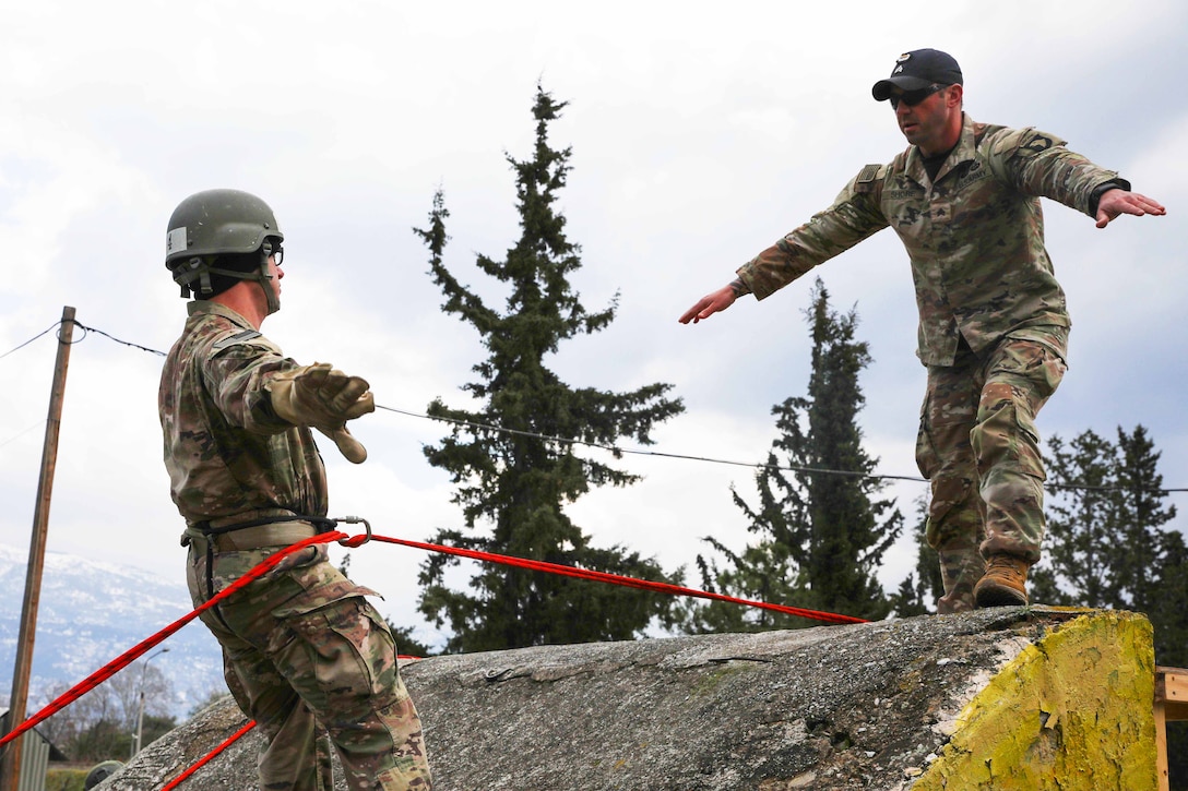 A soldier instructs and demonstrates a position to a Marine wearing a safety helmet and safety rope.