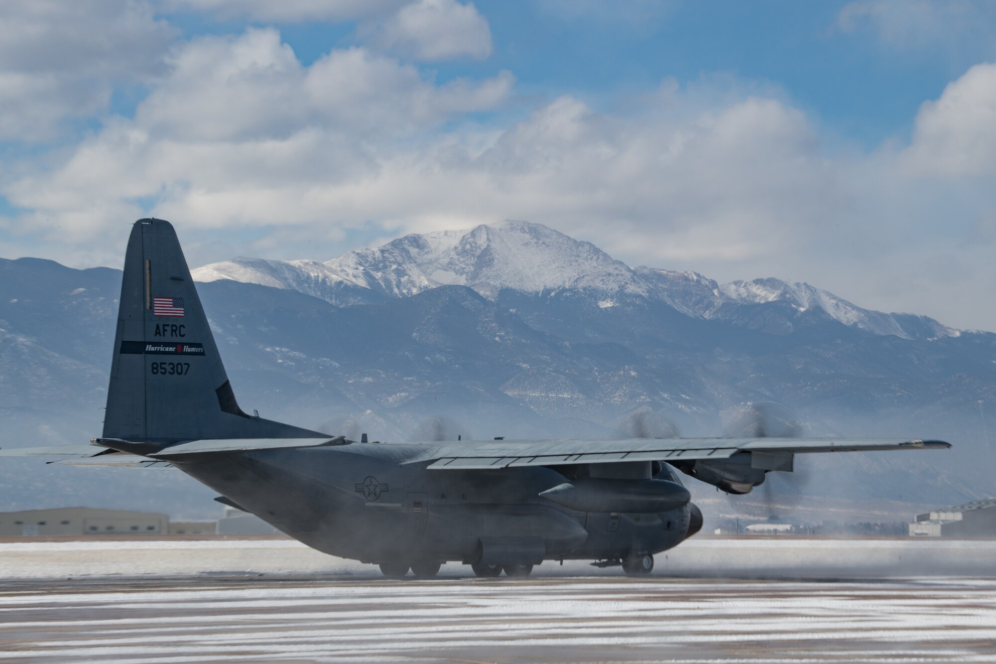 A C-130 with engines running taxis across a snowy flight line with snow-capped Pikes Peak in the distance.