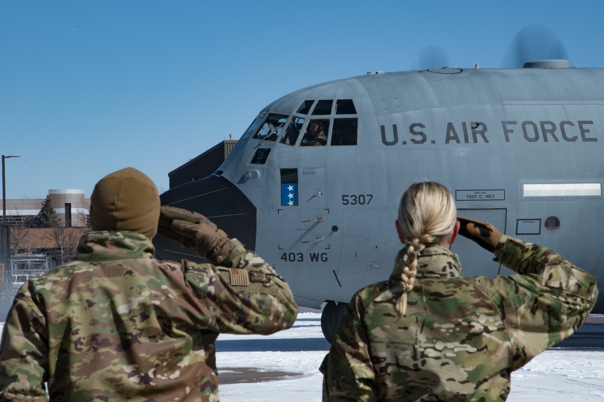 One man and one woman in uniform salute a 403rd Wing C-130 aircraft with 3 stars in the lower window.