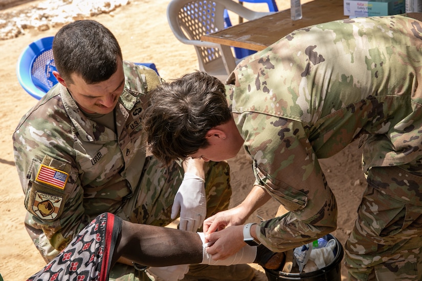 Two people in military uniform administer medical care to a person’s leg.