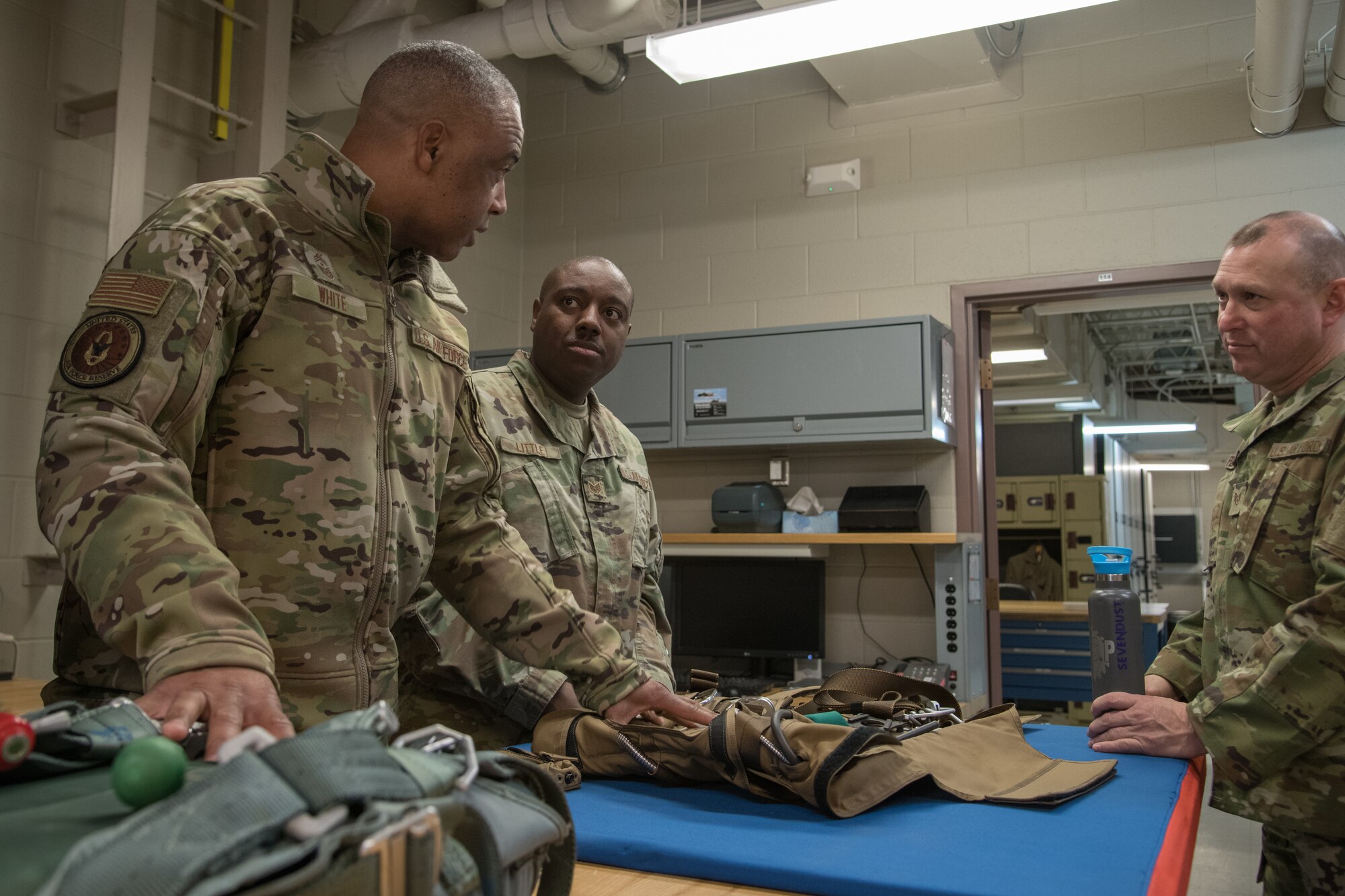 Three men talk while stand around a table with a parachute on a blue mat.