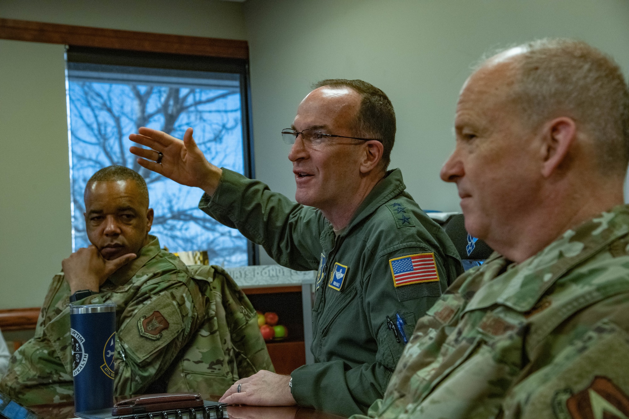 Three men sit at a table, one of them with three stars on the shoulder of his flight suit and his hand in the air while speaking.