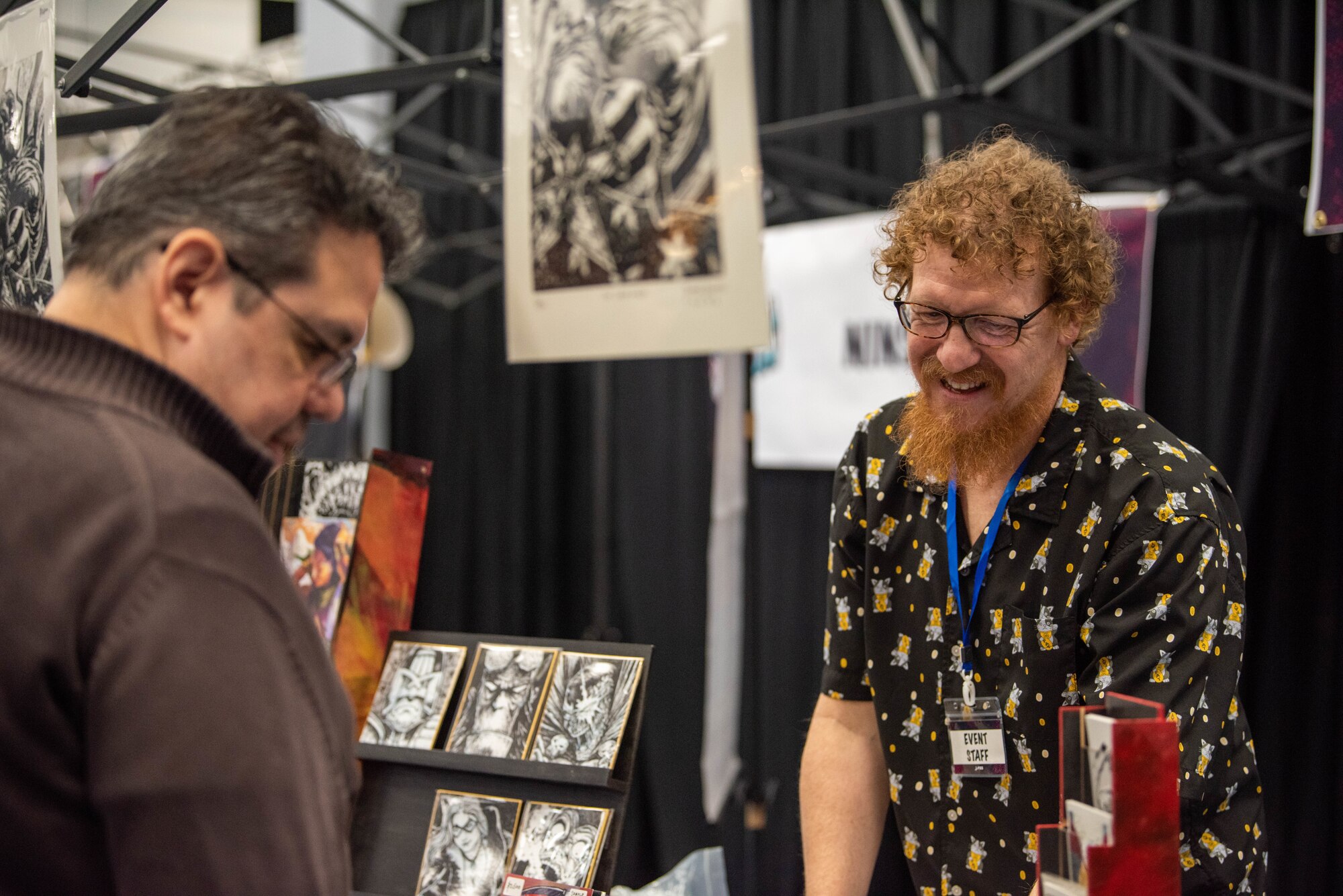 A man with red hair smiles as he stands behind a table displaying artwork.