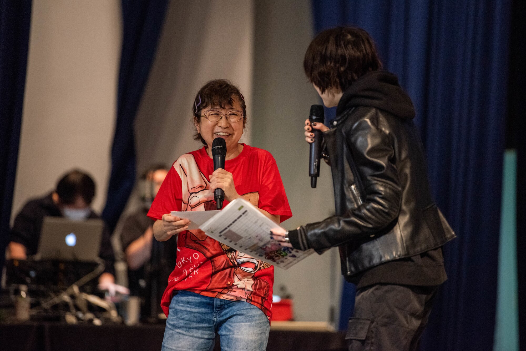 A small, old Japanese woman laughs on a stage.