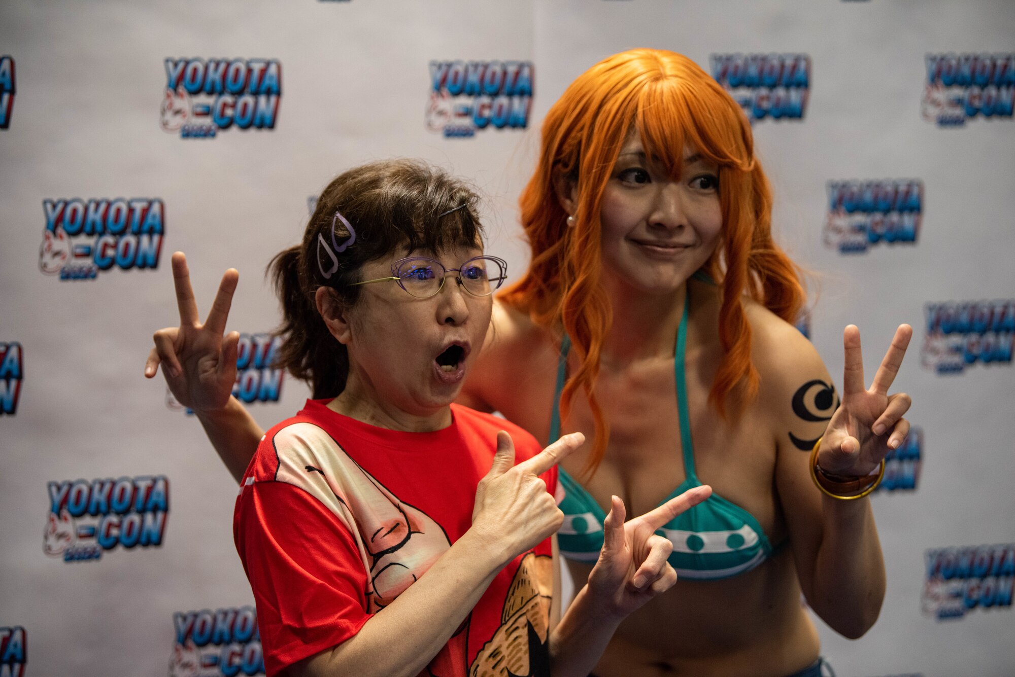 A small, old woman wearing a red shirt stands next to a woman wearing a long red-haired wig and a bikini top.