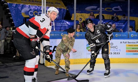 Photo of Sergeant Megan Kennedy ceremonially drops the puck as two hockey players pose as if they are facing off.