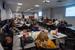 A large group of men and women seated in a classroom setting listening to a dark skinned man in a tan business suit talk about Lean Six Sigma concepts.