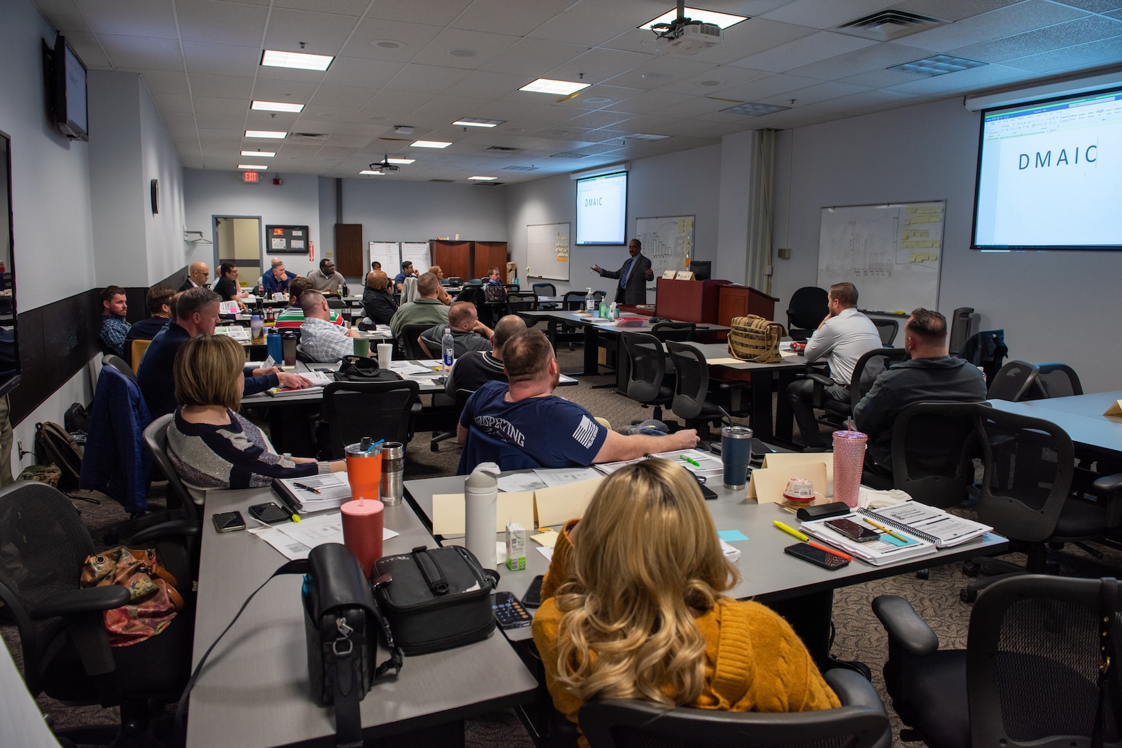 A large group of men and women seated in a classroom setting listening to a dark skinned man in a tan business suit talk about Lean Six Sigma concepts.