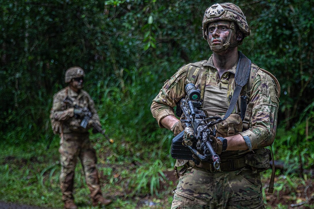 Two soldiers stand, with painted faces, holding weapons in a wooded area.