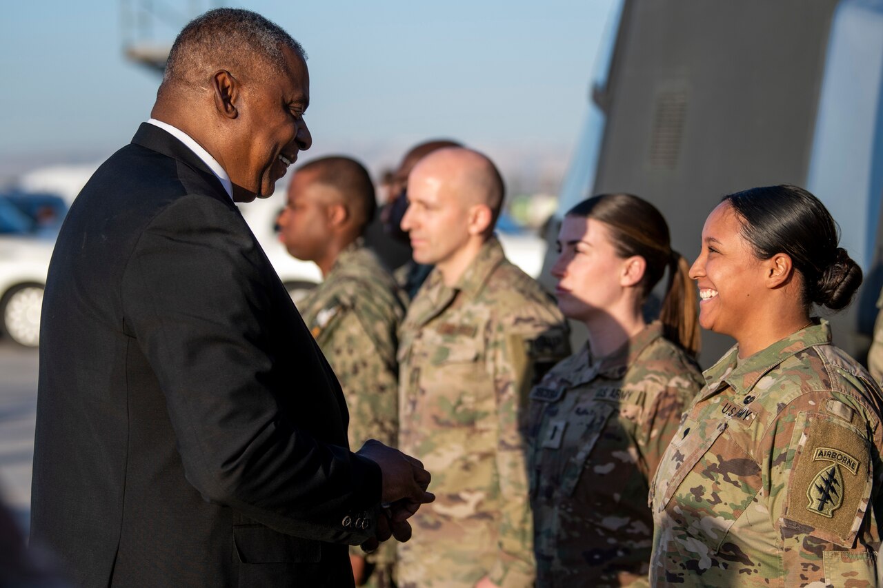 A man in a suit talks with a woman in a military uniform. Many other uniformed personnel stand nearby.