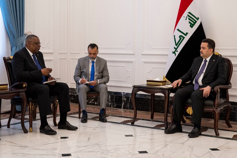 Three men are seated near each other. An Iraqi flag is on display.