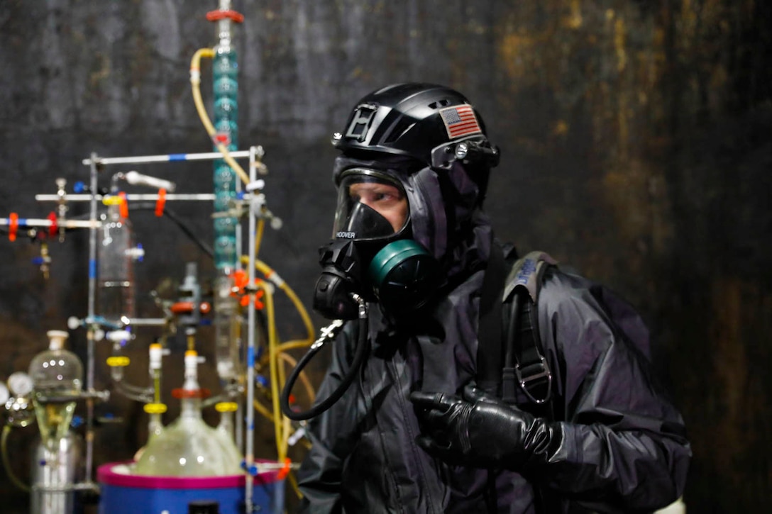 A soldier in protective gear stands by a contraption with tubes and metal bars.