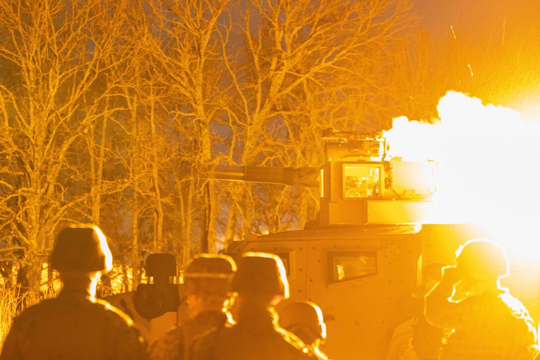 Marines stand next to a tank in the woods as it fires a weapon.