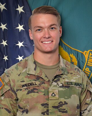 Male Soldier in U.S. Army uniform standing in front of two flags.