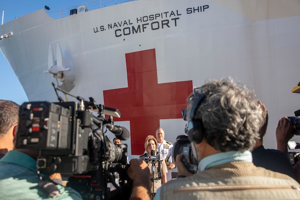 A female soldier speaks into a microphone; a large ship with a red cross painted on the hull is behind her.
