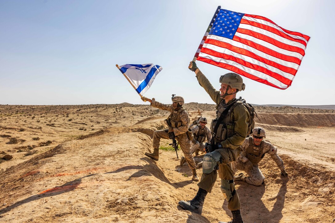 A Marine holds up a U.S. flag and Israeli solder holds one from that nation while on flat, barren terrain.