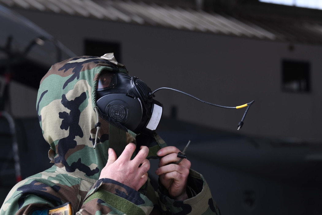A service member wearing a black gas mask puts the hood of his protective camo gear