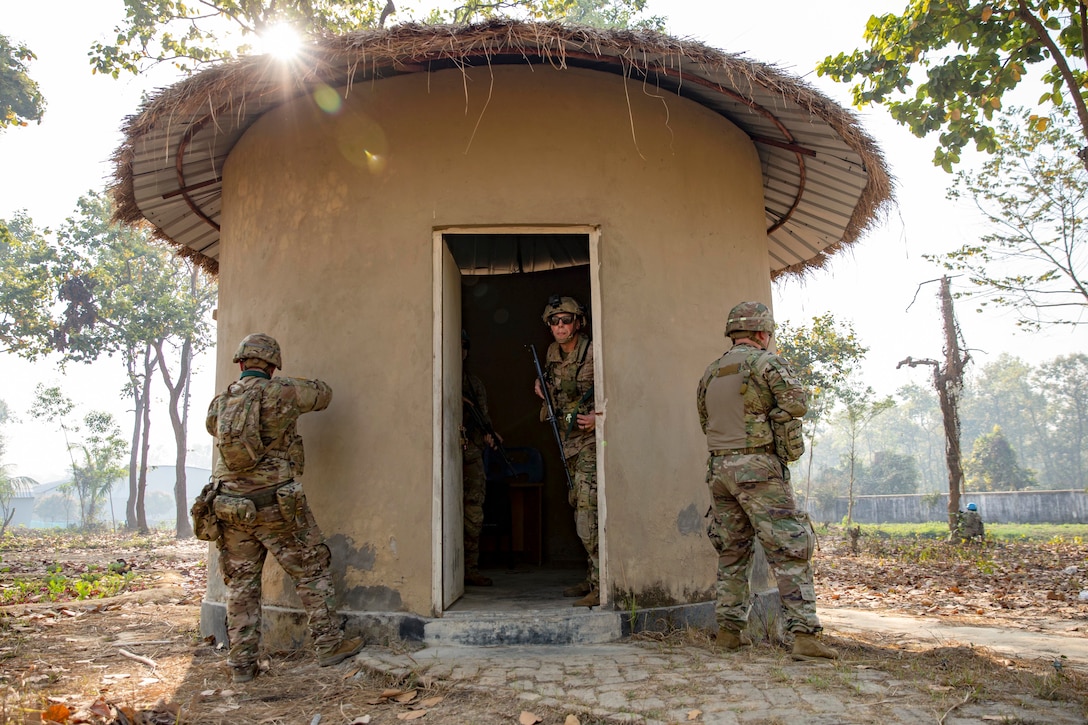 Two guardsmen hold weapons outside of a structure in the woods as  a fellow guardsman exits.