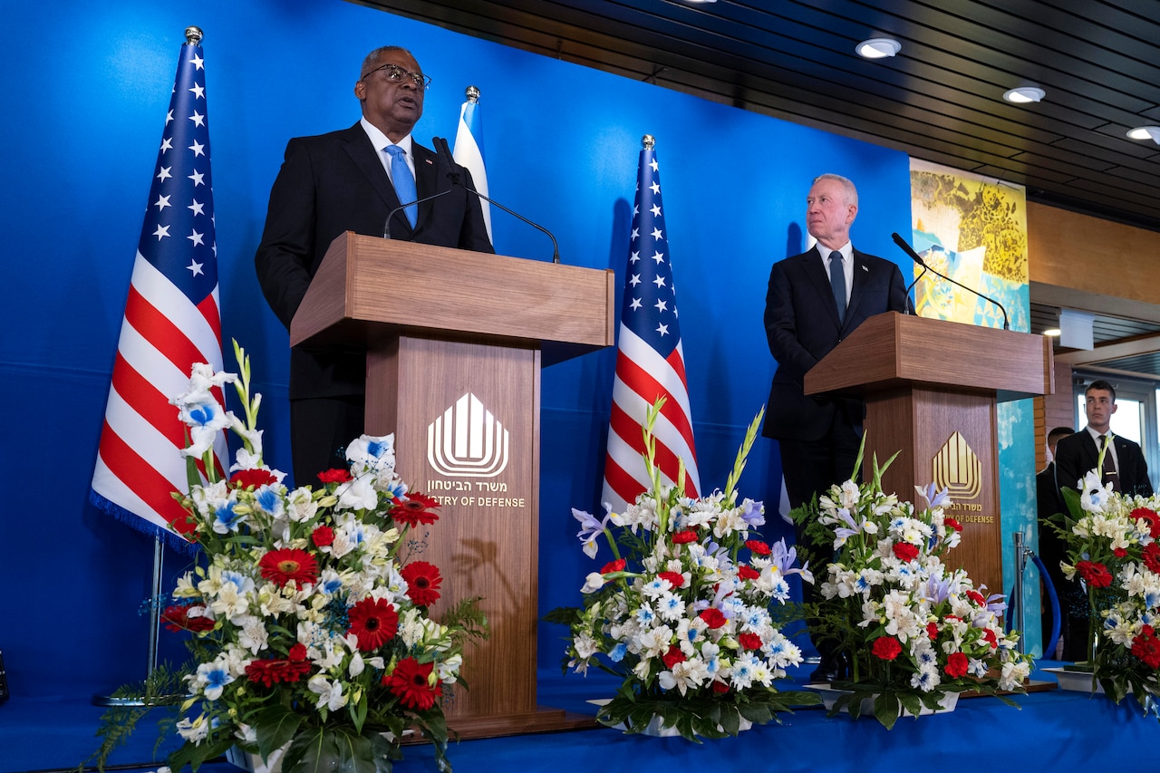 Two men stand on  a stage with flags and flowers and speak to reporters.