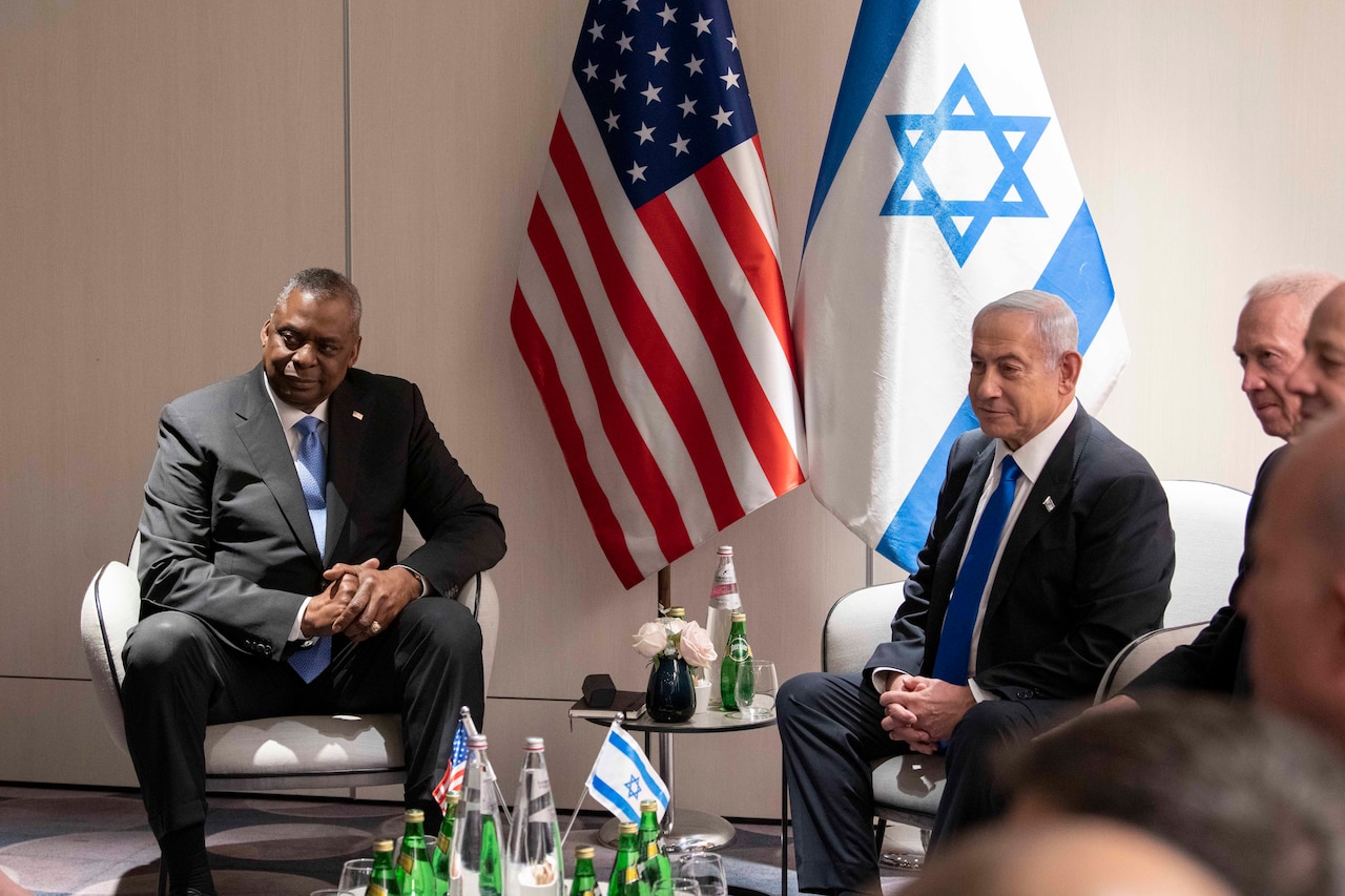 Two men are seated near each other. In the background are the U.S. flag and the flag of Israel.
