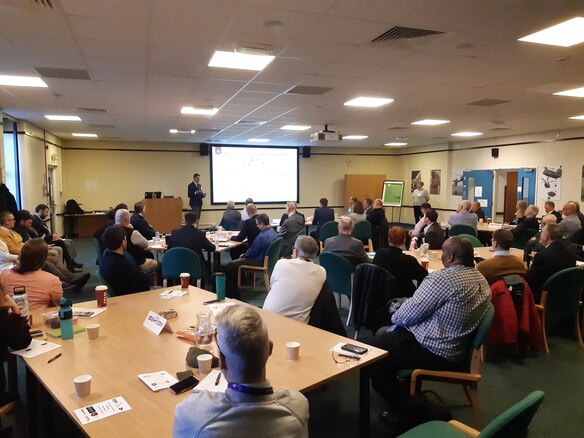 Photo of people sitting at tables listening to a speaker at the front of the room. 