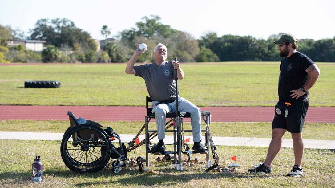 McLeroy's wheelchair sits to his left while he prepares to release the shot from a throwing chair.  At right a coach looks on.