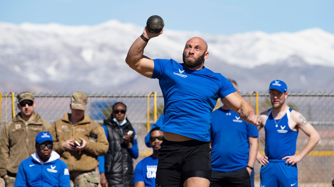 An athlete with snow-covered mountains in the background releases a shot put while other athletes and staff watch from behind.