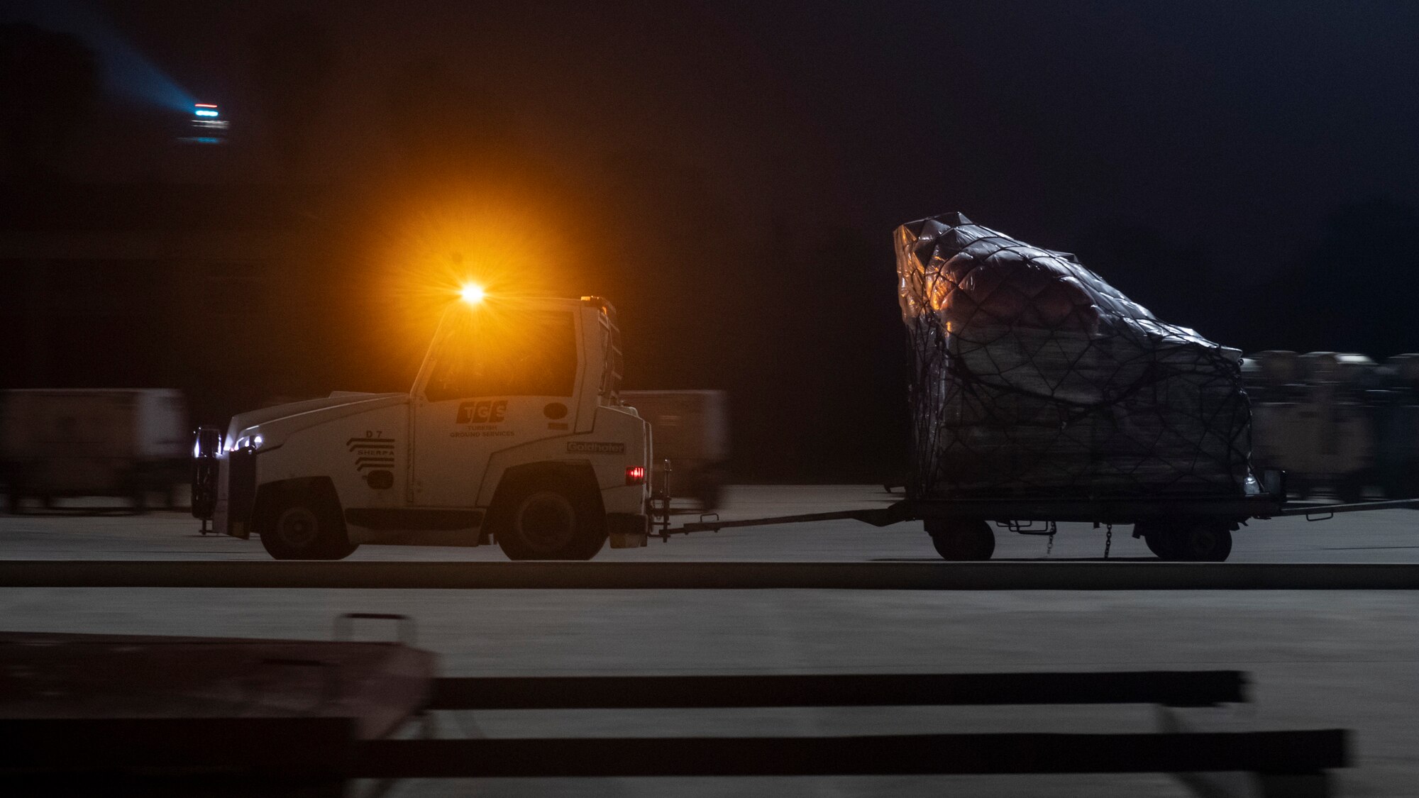 An airport baggage truck hauls humanitarian cargo from the flight line to a transient storage area in support of Turkish government earthquake relief operations March 3, 2023, at Incirlik Air Base, Türkiye. U.S. military organizations are working with interagency colleagues to harness the unique capabilities available to assist those affected by the February 6 earthquakes. The U.S. military’s role during these relief missions is to rapidly respond to this natural disaster with critical support capabilities and life-saving equipment, transporting assistance for aid areas the government of Türkiye deems most necessary. (U.S. Air Force photo by Staff Sgt. Peter Reft)