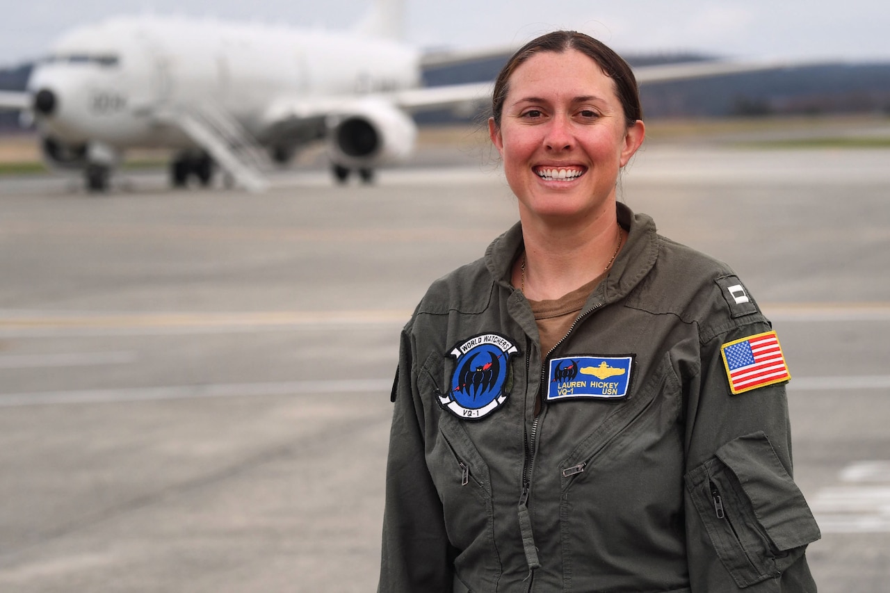 A woman in military uniform stands on a flight line. In the rear is a large aircraft.