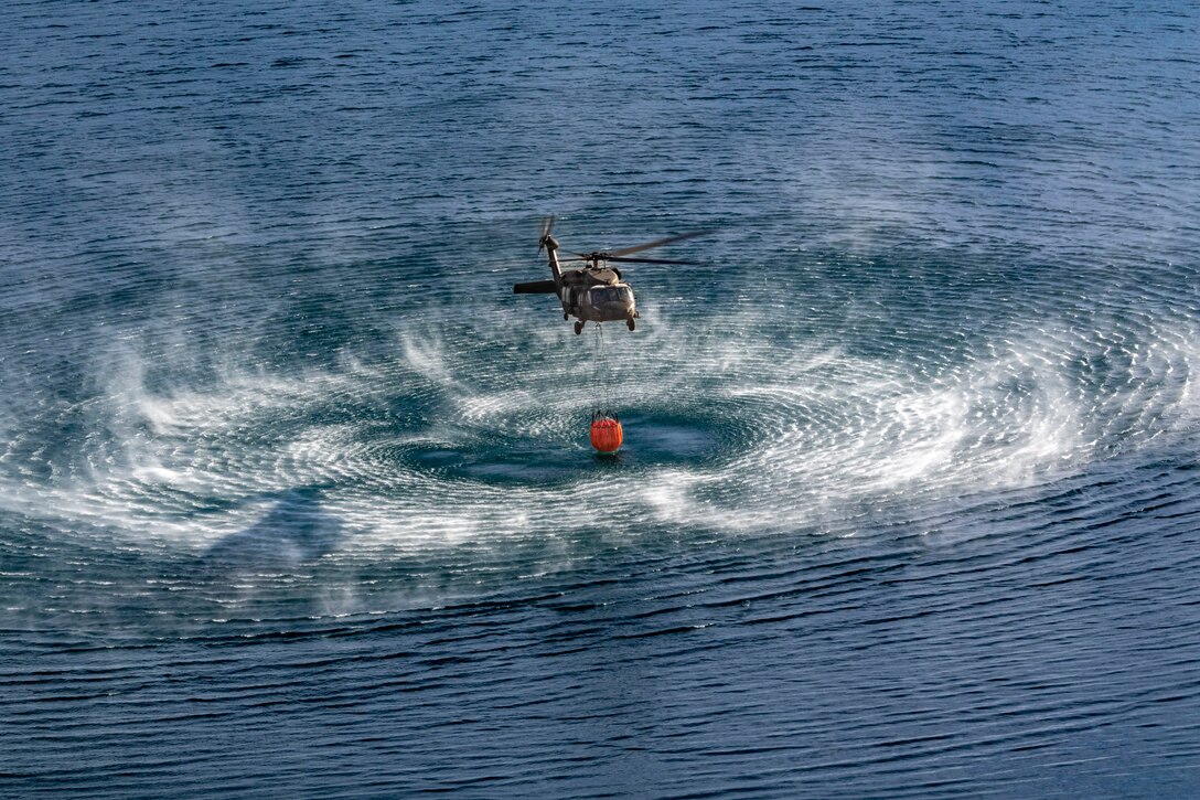 A helicopter creates spray and waves as it flies low over water with a suspended bucket,