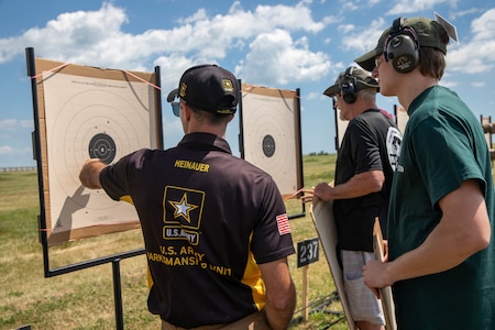 Man in USAMU shooting uniform instructing 2 men on outdoor pistol range.